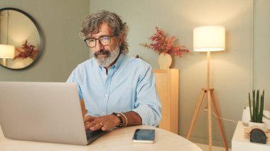 Gri saçlı, mavi tişörtlü yaşlı bir adam modern apartmanın oturma odasında dizüstü bilgisayar kullanarak oturuyor.