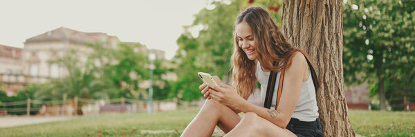 Красивая девушка с длинными волнистыми волосами в белом топе пользуется мобильным телефоном, сидя на зеленой траве под деревом в парке. Девушка флип фотографии на телефоне с пальцами