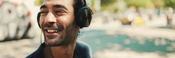 Closeup of young italian guy with ponytail and stubble sits with headphones on street bench and listens to music on cityscape background