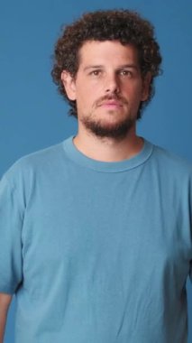 Kollarını kavuşturup kameraya bakan genç bir adam. Stüdyoda mavi arka planda izole edilmiş bir gülümsemeyle.