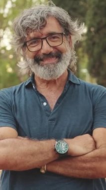 Yaşlı bir adam kollarını kavuşturup gülümseyerek kameraya bakıyor.