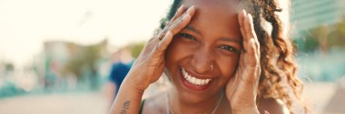 Kentin arka planında uzun kıvırcık saçlı, gülümseyen bir kadının yakın plan portresi. Saçı düzelten ve kameraya bakan mutlu kızın önden yakın çekimi.