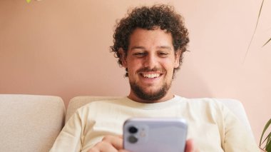 Kıvırcık saçlı, sakallı, rahat oturma odasında koltukta oturan, cep telefonu kullanan pozitif bir genç.