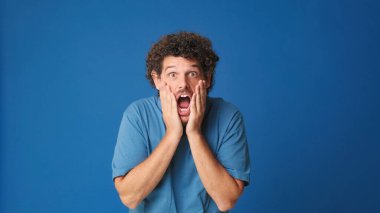 Mavi tişörtlü, kıvırcık saçlı bir adam kameraya büyük gözleriyle bakıyor ve ellerini kaldırıyor. Stüdyonun mavi arka planındaki iyi habere seviniyor.