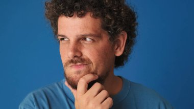 Yakından bakınca, mavi tişörtlü kıvırcık saçlı adam zor bir seçim düşünüyor, aklında bir plan kuruyor, stüdyoda mavi arka planda izole edilmiş bir plan.