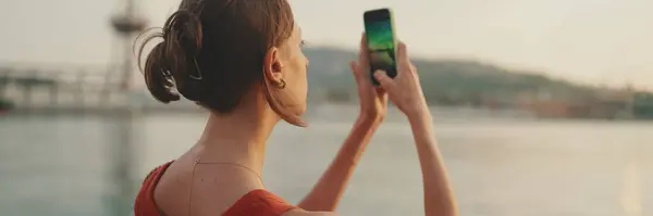 Close-up, girl stands on the embankment and takes pictures of sunrise on a mobile phone. Closeup of young woman shoots the sea on smartphone in the morning time. Back view