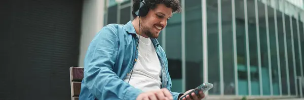Close-up of young bearded man in denim shirt sitting in headphones on bench listening to music and using mobile phone on modern buildings background