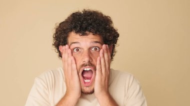 Yakından, bej tişörtlü, şaşkın adam kameraya büyük gözlerle bakıyor, ellerini yüzüne kaldırıyor, vay be, stüdyoda bej arka planda izole edilmiş.