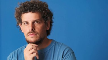 Yakından, mavi tişörtlü kıvırcık saçlı adam, problemini düşünüyor, stüdyoda mavi arka planda izole edilmiş bir şey hatırlıyor.