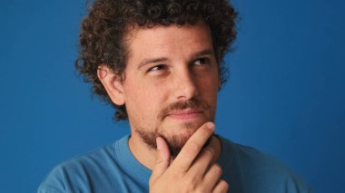 Yakından bakınca, mavi tişörtlü kıvırcık saçlı adam zor bir seçim düşünüyor, aklında bir plan kuruyor, stüdyoda mavi arka planda izole edilmiş bir plan.