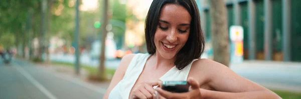 Beautiful woman with freckles and dark loose hair wearing white top sits on bench with phone in her hands. Cute smiling girl watching photo, video on mobile phone modern city background