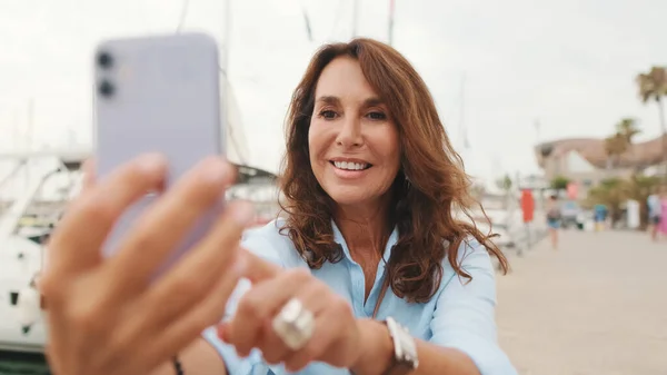Beautiful smiling slim middle-aged woman sits in the port on yacht background uses mobile phone, watches funny videos, chats with friends on social networks