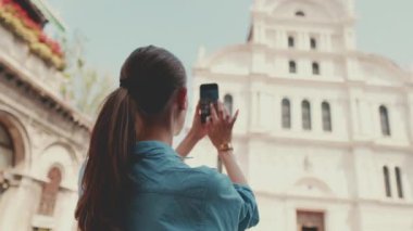 Uzun kahverengi saçlı, mavi gömlekli güzel bir kız eski şehrin meydanında dikilirken cep telefonuyla fotoğraf çekiyor.