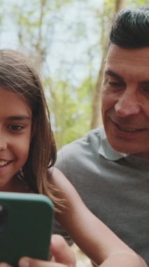 Yakın çekim, mutlu aile akıllı telefon kullanarak parktaki bankta oturan resimlere ve videolara bakıyor.