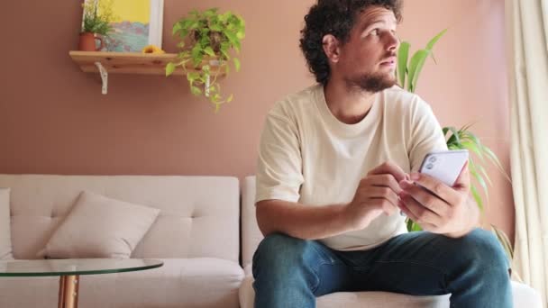 Guy Senta Sofá Acolhedora Sala Estar Segurando Telefone Suas Mãos — Vídeo de Stock