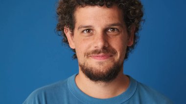 Mavi tişörtlü kıvırcık saçlı bir adam stüdyoda mavi arka planda gülümseyen bir kameraya bakıyor.