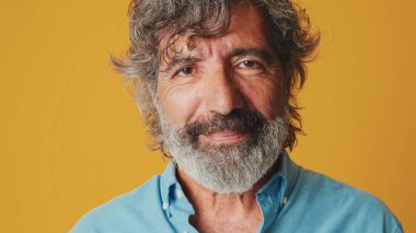 Yakın plan portre, mavi gömlek giyen yaşlı gri sakallı adam, kameraya gülümseyerek bakıyor, stüdyonun turuncu arka planında izole edilmiş.