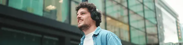 Emotional young bearded man in denim shirt walks down the street in headphones listening to music and sings along, holding mobile phone in his hand on modern cityscape background
