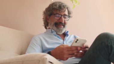 Yaşlı bir adam, elinde telefonuyla oturma odasında kanepede oturuyor.