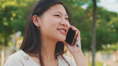 Yakından, neşeli, güzel esmer kız şehir parkında bankta oturuyor, cep telefonuyla konuşuyor.