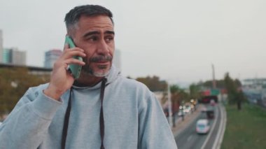 Orta yaşlı spor giyimli bir adam köprüde dururken cep telefonuyla konuşuyor.