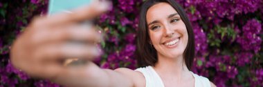 Çilli ve koyu renk saçlı güzel bir kadın beyaz bluz giyerek selfie çekiyor. Şirin gülümseyen kız, çiçek arkaplanda selfie çekiyor.