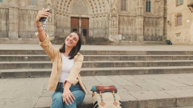 Gezgin kız, cep telefonuyla selfie çekiyor, eski bir Avrupa şehrinin tarihi bölümünde eski bir binanın merdivenlerinde otururken zafer işareti gösteriyor.