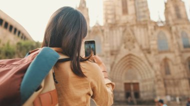 Omuzlarında sırt çantasıyla gezgin bir kız Avrupa 'nın tarihi bölgesinde, arka manzaralı eski bir binanın cep telefonuyla fotoğraf çekiyor.