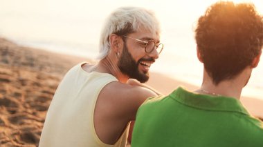 Kapatın, güzel eşcinsel çift şafak vakti kumsalda otururken konuşuyor ve gülüyor.