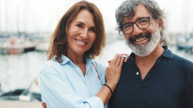 Yaklaş, mutlu yaşlı çift yatın arka planında dikilirken birbirlerine sarılıyorlar başlarını çevirip gülümseyerek kameraya bakıyorlar.