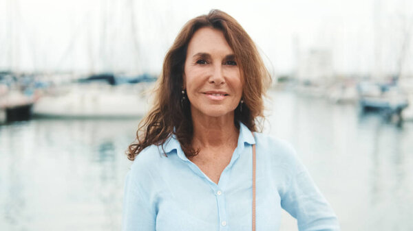 Закрытая, счастливая привлекательная женщина средних лет, стоящая в порту на фоне яхты, с улыбкой смотрит в камеру