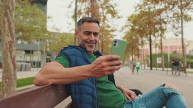 Orta yaşlı bir adam parkta bankta oturuyor ve cep telefonu kullanıyor.