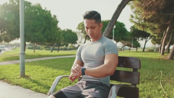 年轻人坐在公园的长椅上 用的是聪明的手表 — 图库视频影像