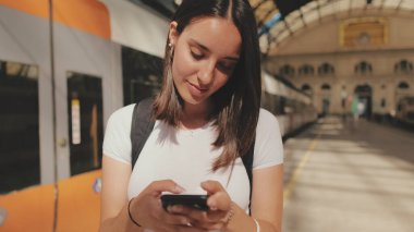 Güzel kız gezgin, tren istasyonunun platformunda dururken cep telefonu kullanıyor.