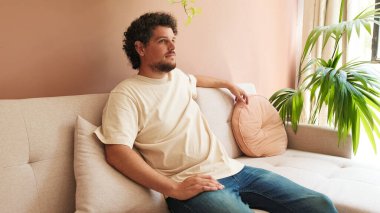 Kıvırcık saçlı ve sakallı genç adam rahat oturma odasında kanepede dinleniyor.