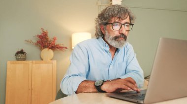 Gri saçlı, gözlüklü, yorgun yaşlı bir adam modern dairede oturma odasında oturmuş dizüstü bilgisayarla çalışıyor.