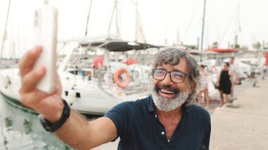 Yakın plan, gözlüklü mutlu orta yaşlı adam yat arka planında oturur, akıllı telefon kullanır, cep telefonunda selfie çeker.