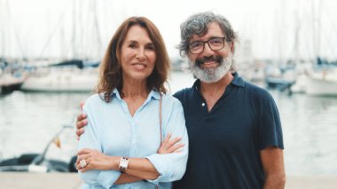 Yaklaş, mutlu yaşlı çift yatın arka planında dikilirken birbirlerine sarılıyorlar başlarını çevirip gülümseyerek kameraya bakıyorlar.