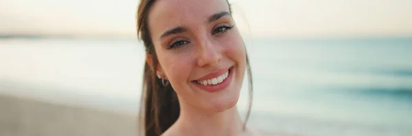 stock image Young athletic woman with long ponytail wearing beige sports top smiling makes selfie on the sea background