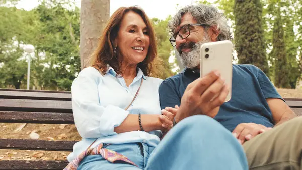 stock image Retired couple talking, having fun using mobile phone while sitting outside in the park in autumn