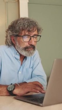 Gözlüklü, orta yaşlı bir adam oturma odasında oturmuş dizüstü bilgisayarla çalışıyor.