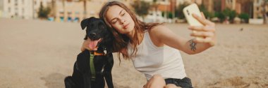 Öğle yemeği vakti. Şirin kız kumsalda evcil hayvanlı sandviçiyle oturuyor. Kız köpeğine sandviçle davranıyor. Modern şehir arka planında sahilde öğle yemeği.