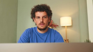 Oturma odasında oturan kıvırcık saçlı ve sakallı genç adam dizüstü bilgisayarına bakıyor.