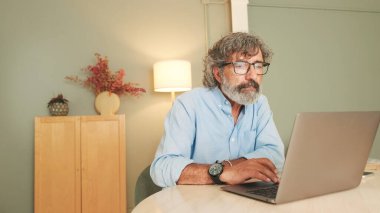 Gri saçlı, gözlüklü, düşünceli yaşlı bir adam modern dairede oturma odasında oturmuş dizüstü bilgisayarla çalışıyor.