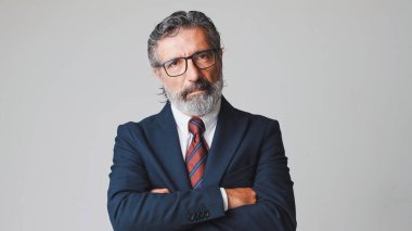 Gözlüklü, gri saçlı, kollarını kavuşturup kameraya bakan orta yaşlı bir işadamı. Stüdyonun gri arka planında izole edilmiş.