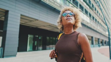 Gözlüklü, kahverengi elbiseli, sırtında sırt çantasıyla genç bir kadın modern binalarda sokakta yürüyor.