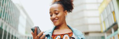 At kuyruklu tatlı Afrikalı kız, kot ceket giyiyor, ulusal desenli ekin giyiniyor, elinde telefonla modern binaların arka planında yürüyor..