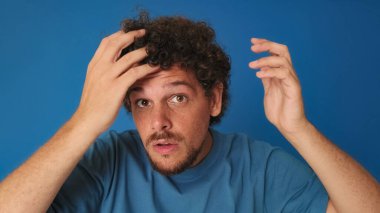 Mavi tişörtlü, kıvırcık saçlı, aynaya bakar gibi kameraya bakan stüdyoda saçını mavi arka planda düzelten adamın yakın çekimi.