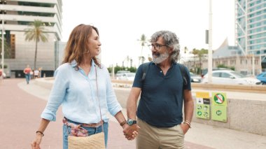 Orta yaşlı evli çift yaya geçidinde karşıdan karşıya geçiyor, el ele tutuşuyorlar ve konuşuyorlar.