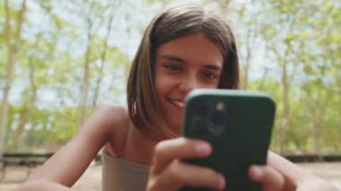 Kapalı, gülümseyen genç kız parkta otururken cep telefonu kullanıyor.